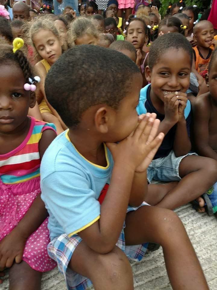 Children sitting together outdoors, smiling and clapping among a crowd