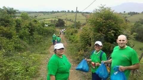 Volunteers in green shirts walk a rural path carrying blue bags under an overcast sky