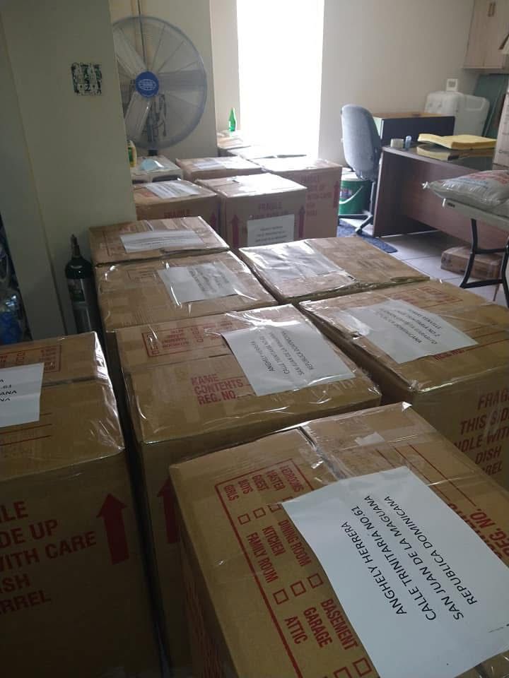 Boxes stacked in a room with a standing fan and desks by a bright window