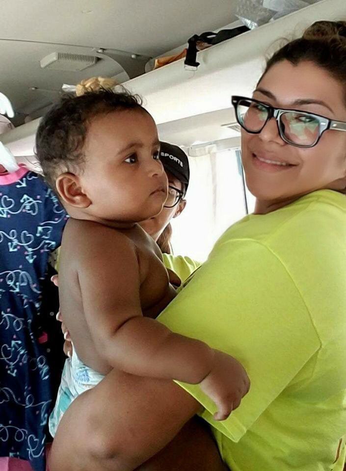 Adult holding a baby indoors, both looking at the camera; woman wears glasses and a neon green shirt.