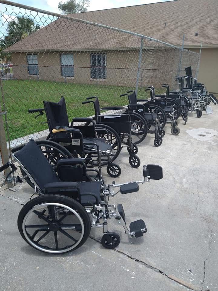 Several wheelchairs lined up on a concrete path beside a brick building and chain-link fence.