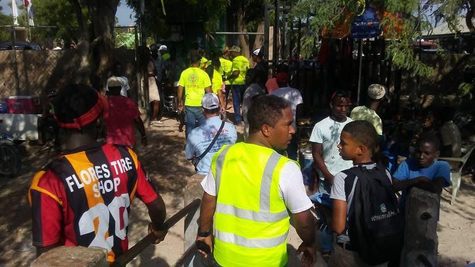 People in bright safety vests guide a crowd outdoors near a gate, with children nearby.