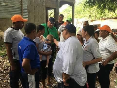 Group of people gathered outdoors, talking around a baby under a shaded structure.
