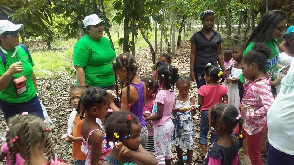 People gathered outdoors in a grove, with adults talking and children standing in a group.