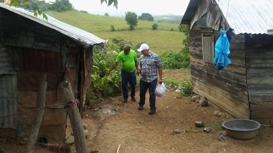 Two people walking between rustic wooden huts on a muddy rural path, carrying bags.