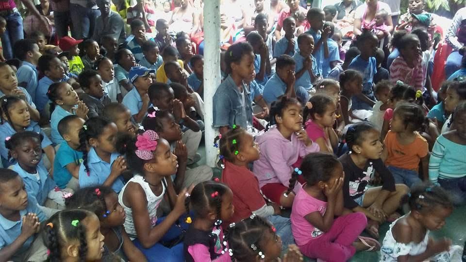 Crowded indoor gathering of children seated closely together, many in colorful clothes, facing the same direction