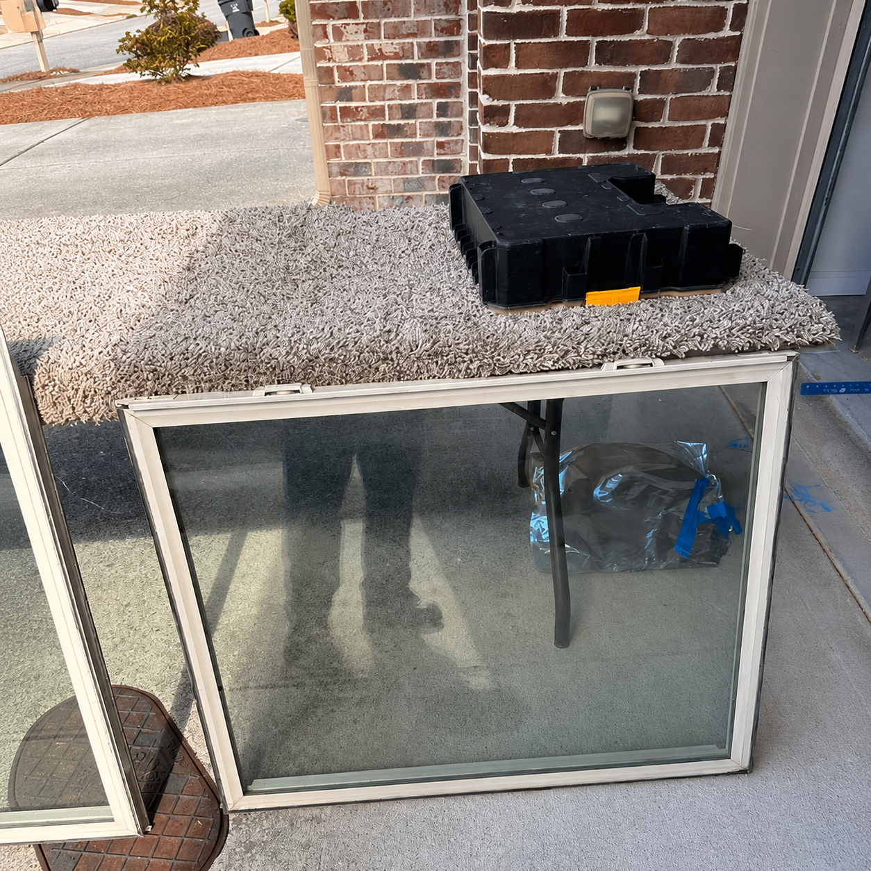 Glass door leaning against a gravel-covered utility box beside a brick wall, with a black toolbox on top.