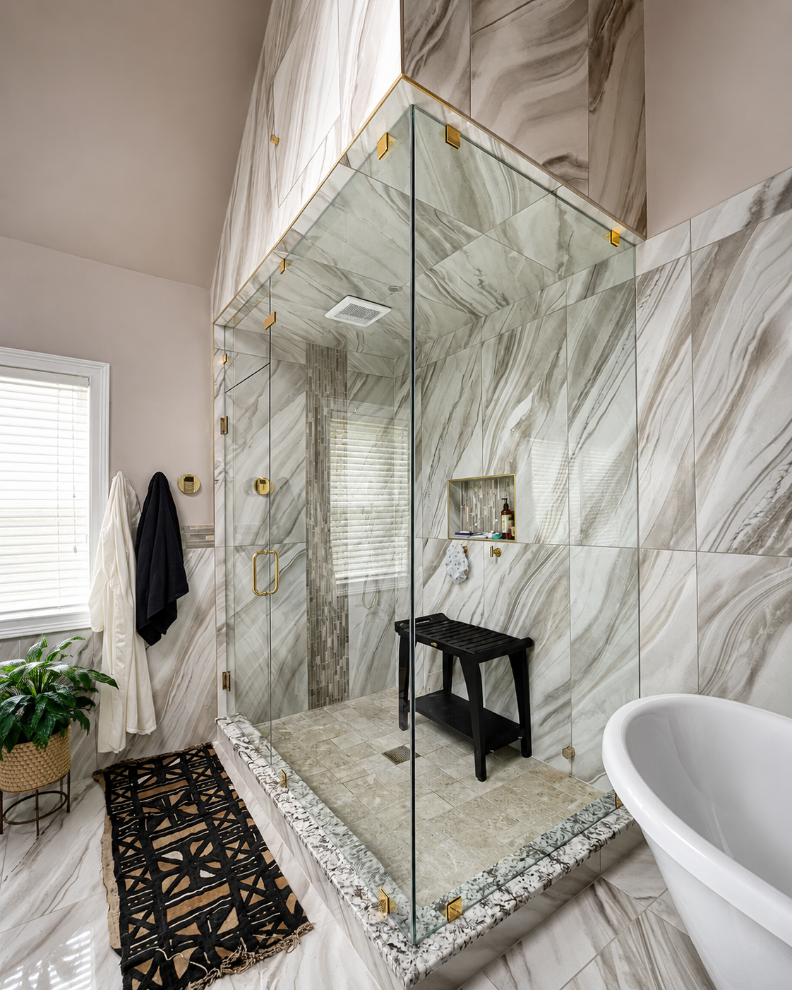 Glass walk-in shower with marble walls, brass fixtures, and a bench in a bright bathroom