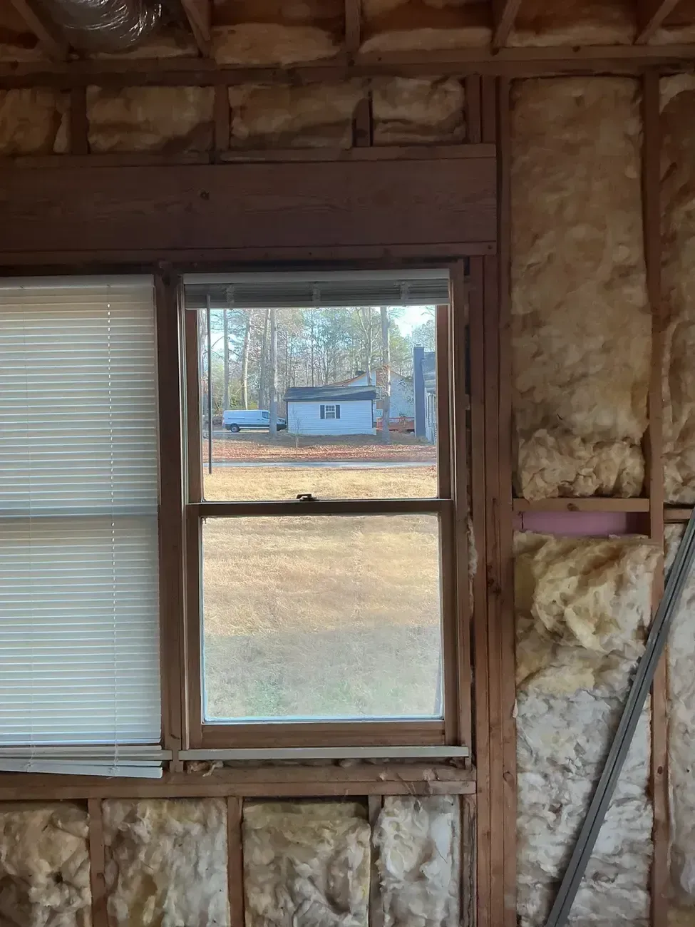 Interior wall framing with insulation and a window overlooking a yard and small blue shed