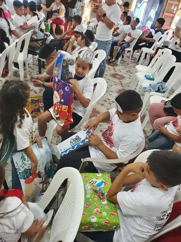 Children seated in rows at a festive indoor gathering, holding colorful wrapped gifts.