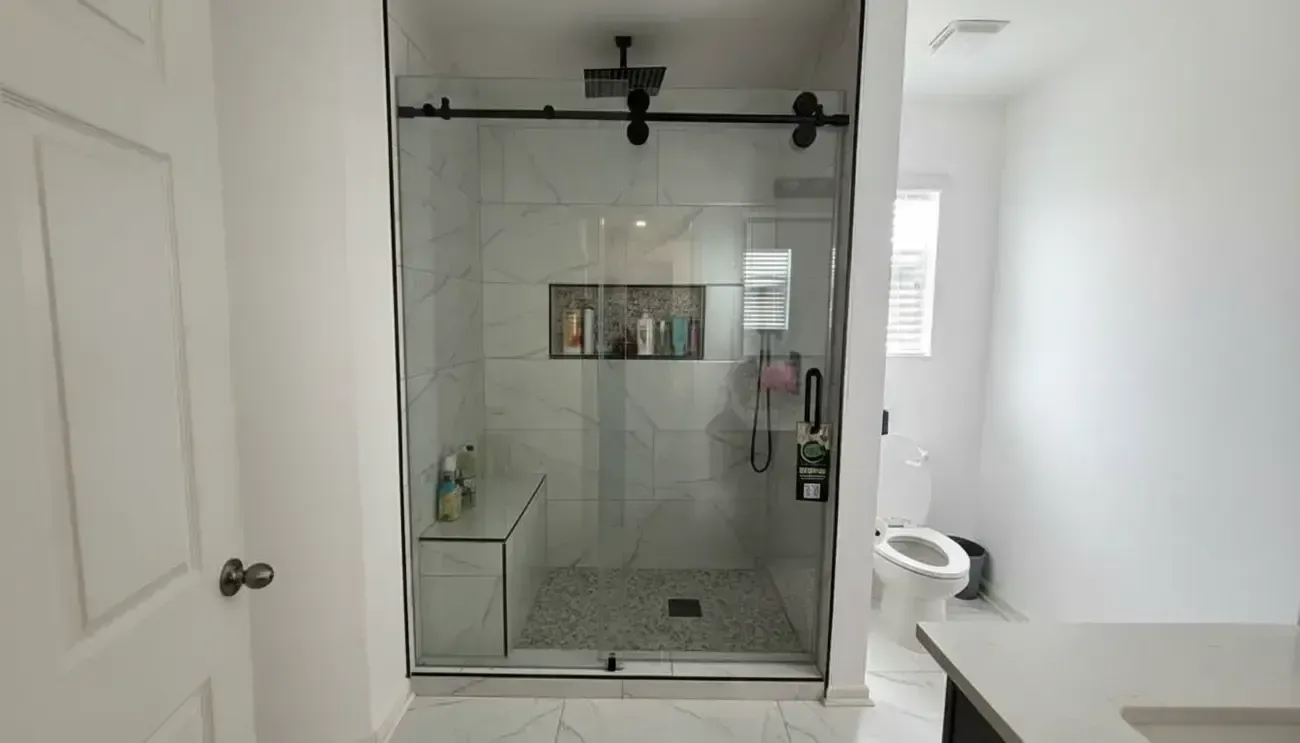 Glass-enclosed shower with black fixtures in a bright white bathroom, with a toilet and sink visible on the right.