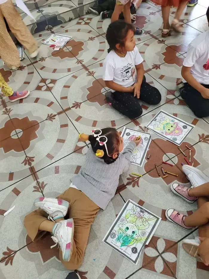 Children sitting on a tiled floor, doing a coloring activity with worksheets and markers.
