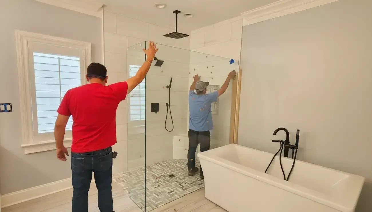 Two workers paint and install a shower wall in a bright bathroom, with a freestanding tub nearby.
