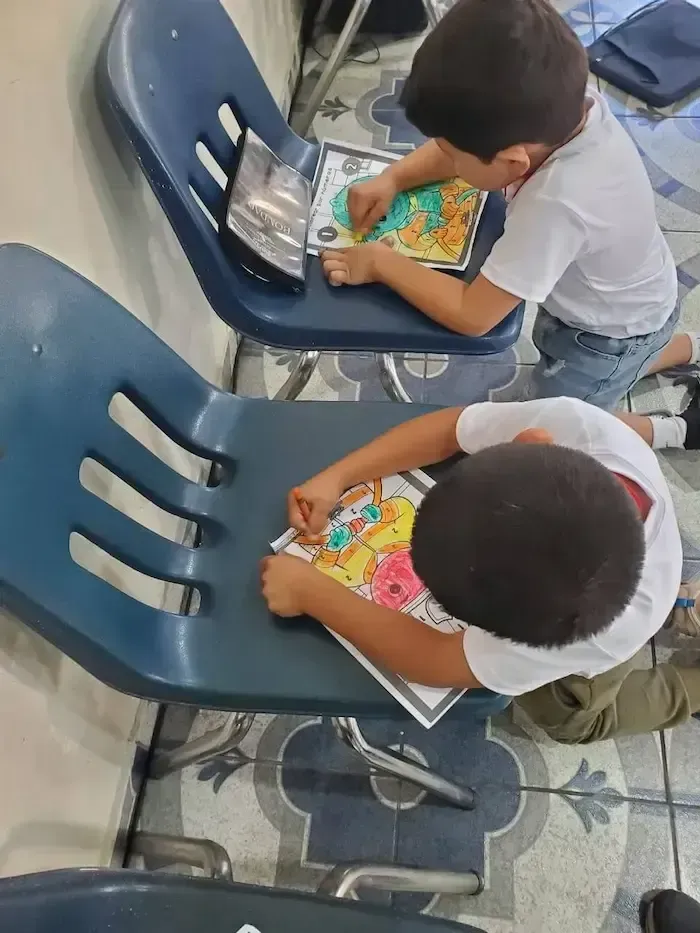Two children lean over colorful workbooks while seated on blue chairs in a classroom.