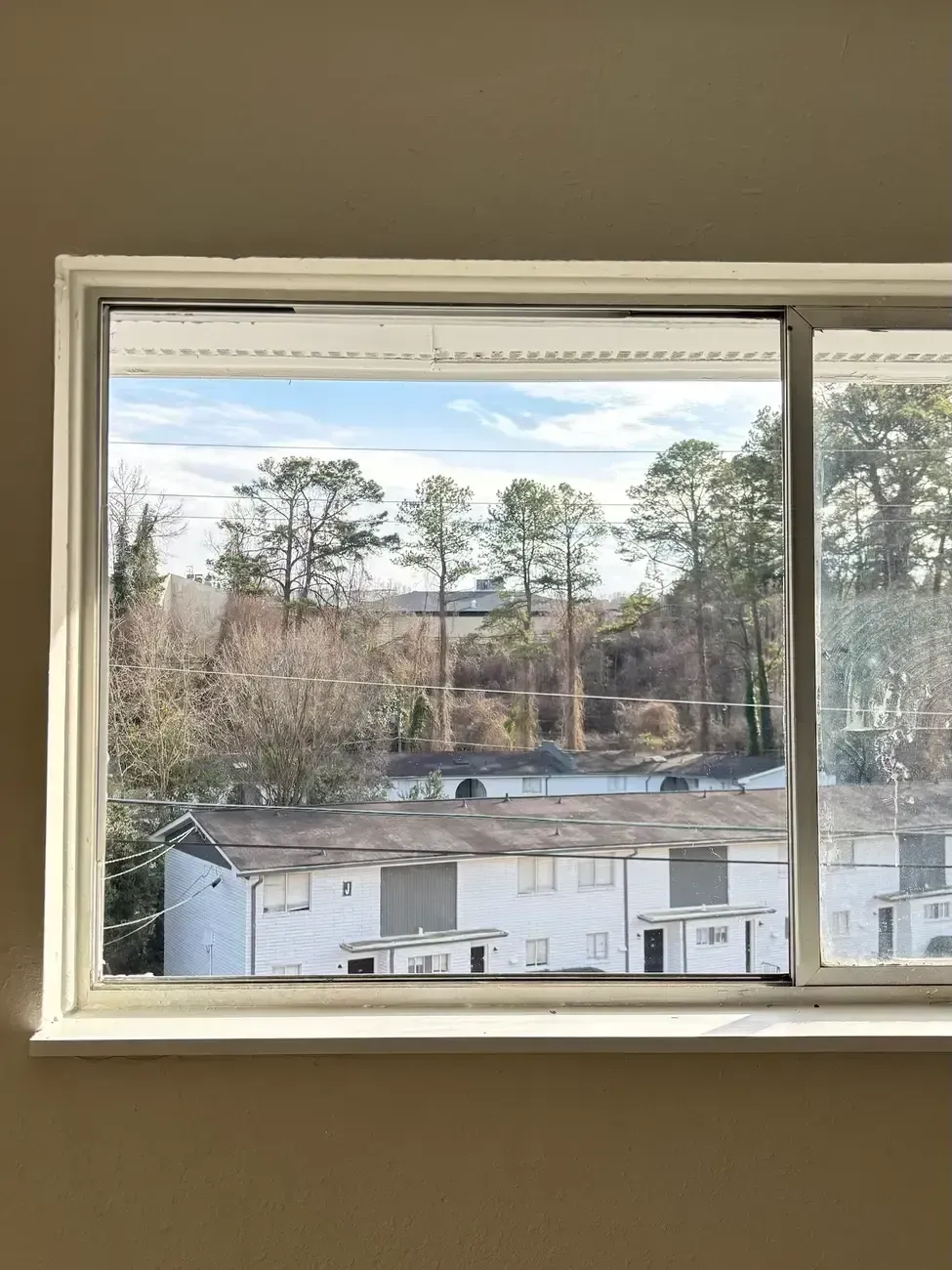 View through a window of a white building, trees, and a cloudy sky.