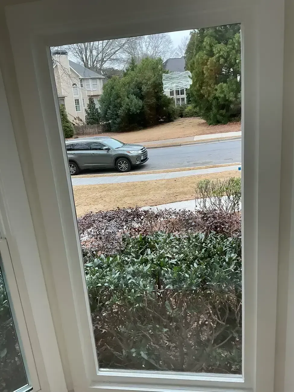 View through a window of a suburban street with a parked gray car, hedges, and houses across the road