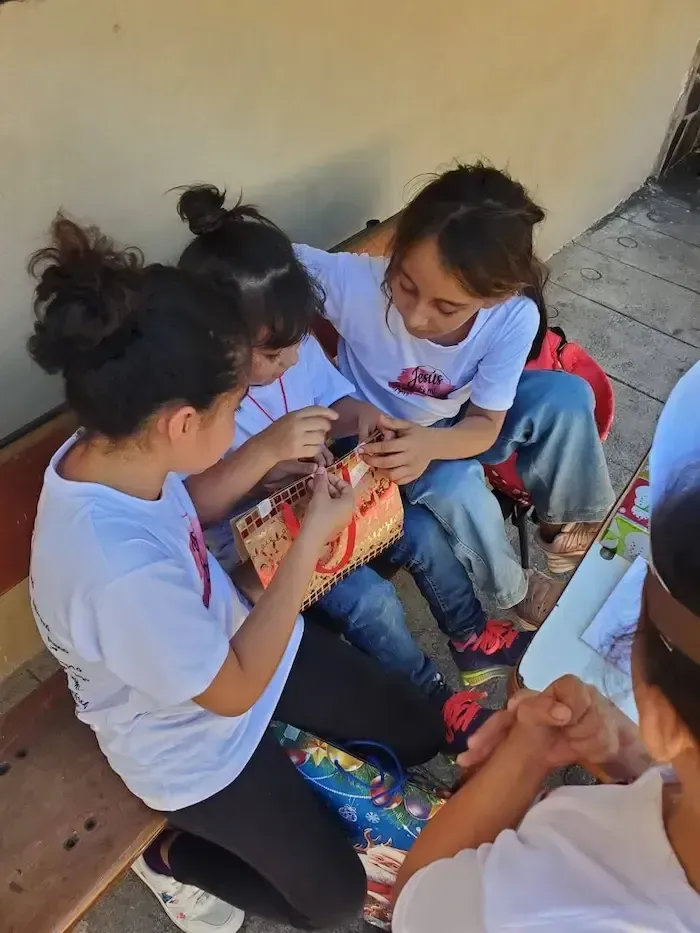 Several children sit together on a bench, chatting and looking at a small object outdoors.