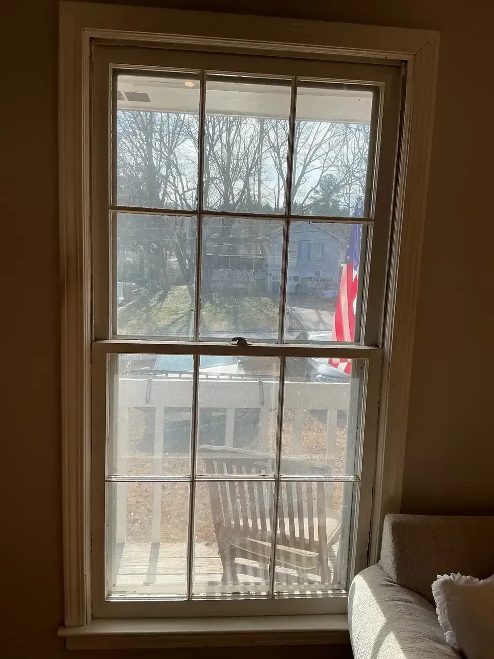 Window with divided panes looking onto a porch and trees, with sunlight and an American flag outside.
