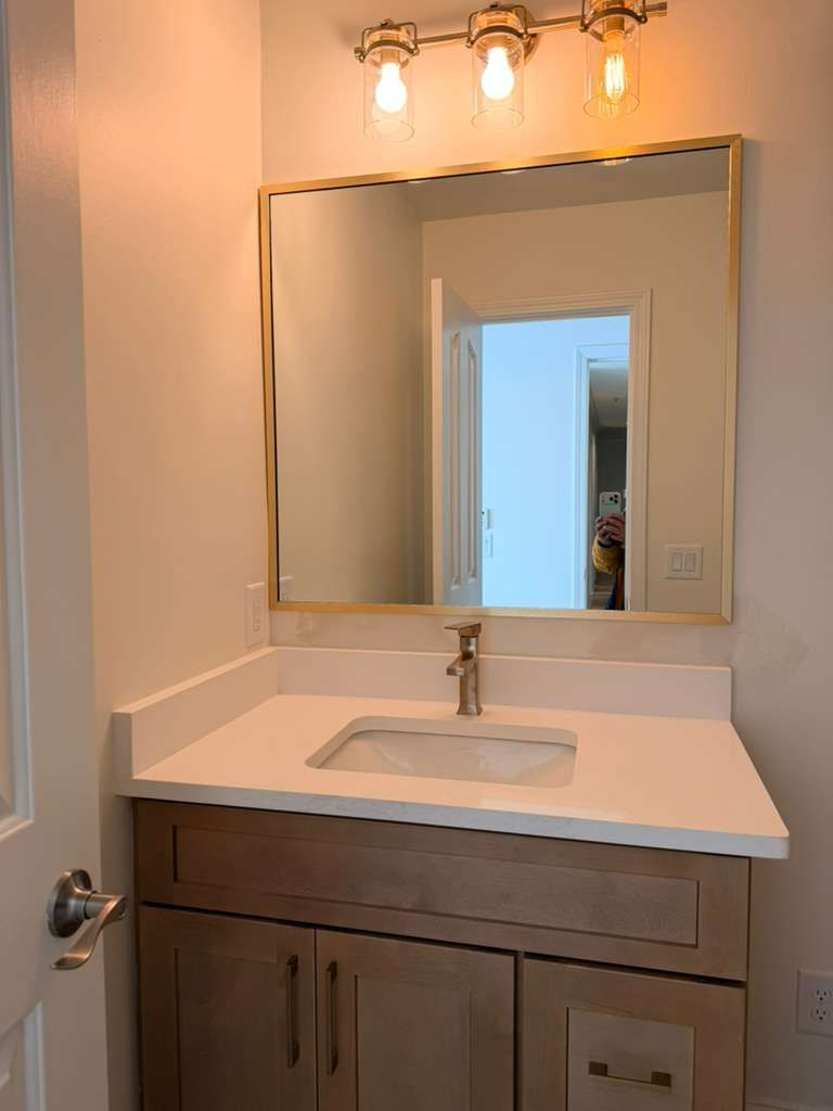Bathroom vanity with white countertop, large mirror, and warm light fixture above a wooden cabinet.