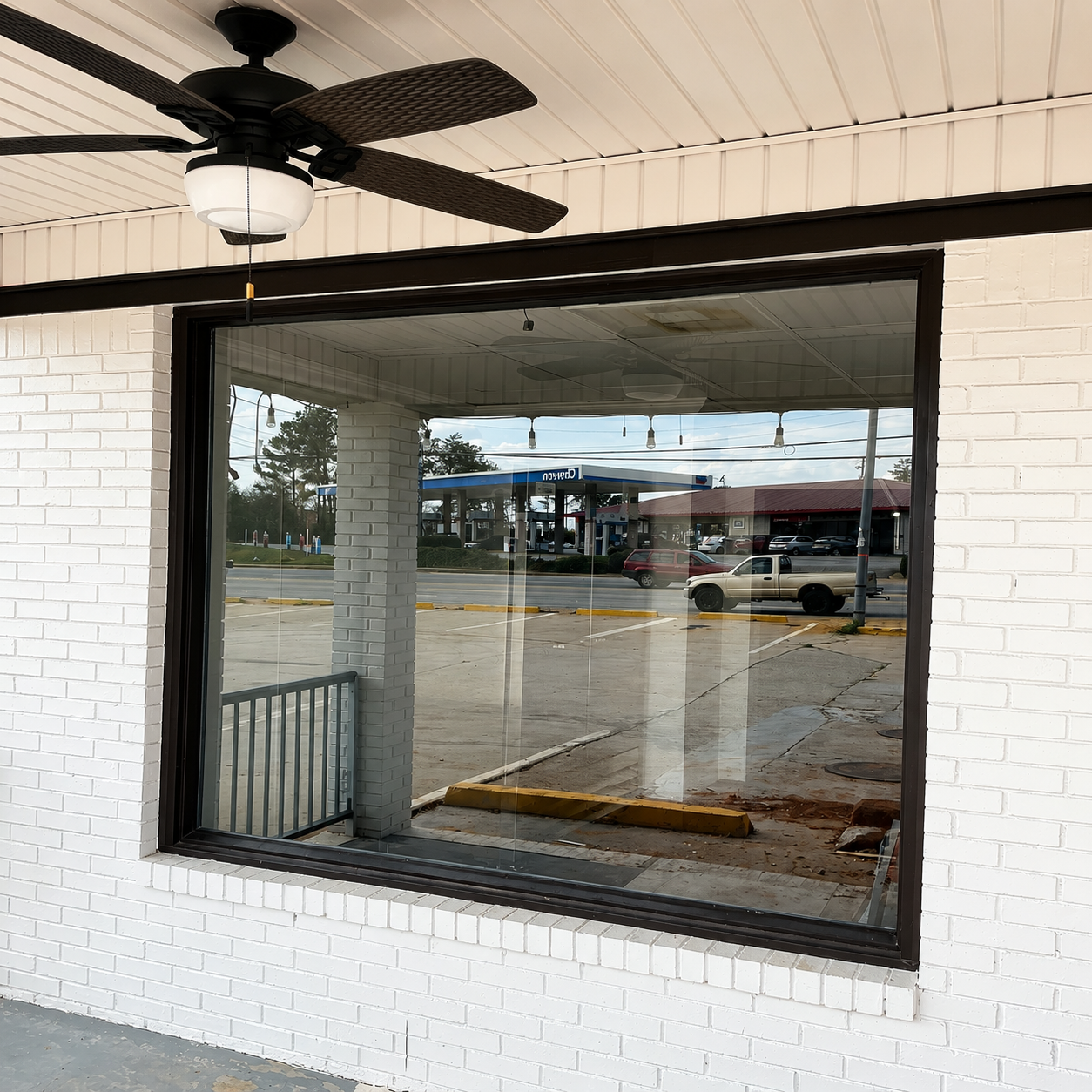 Large window in a white brick wall reflecting a parking lot, with a ceiling fan under the overhang.
