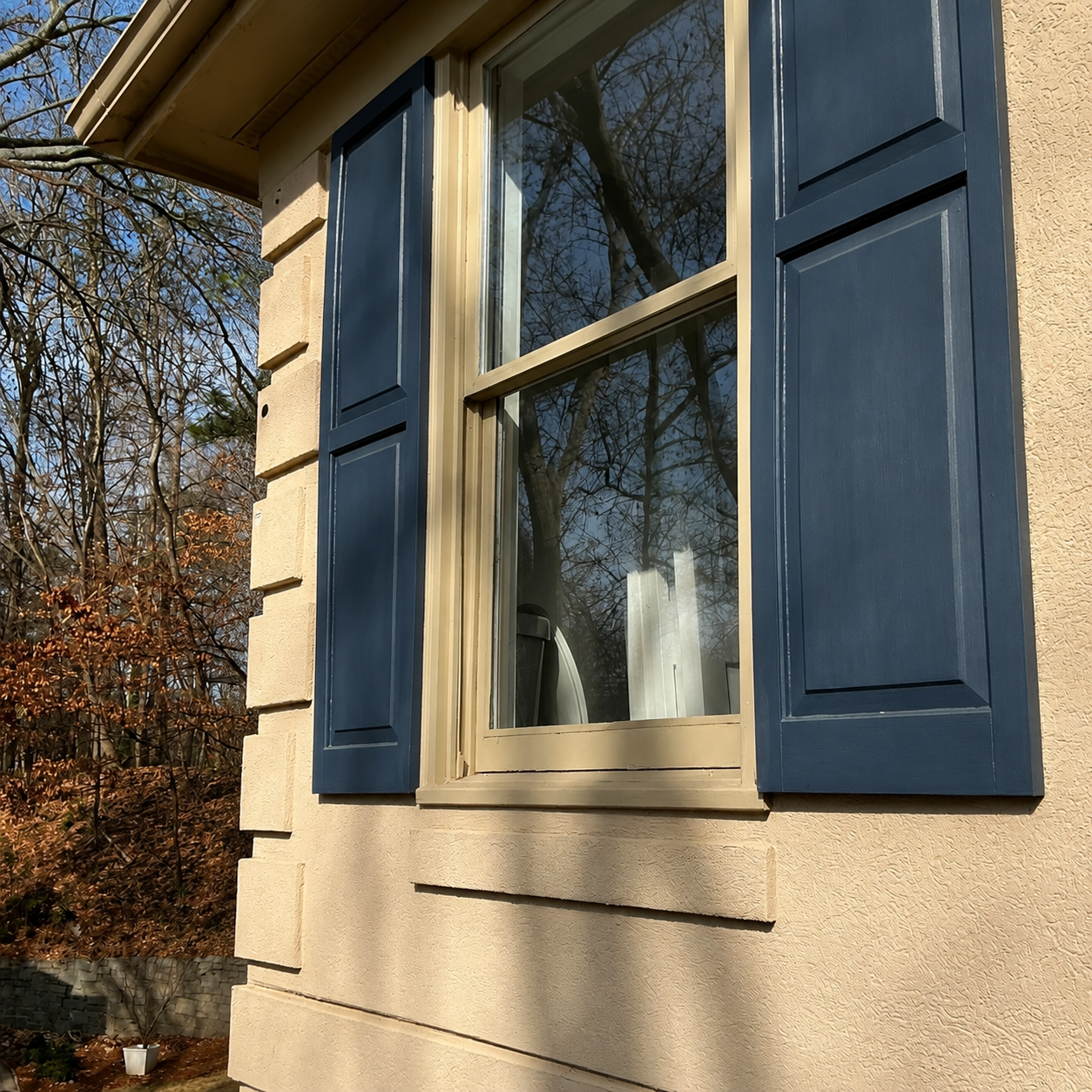 Blue shutters flanking a beige house window with reflections of trees outdoors