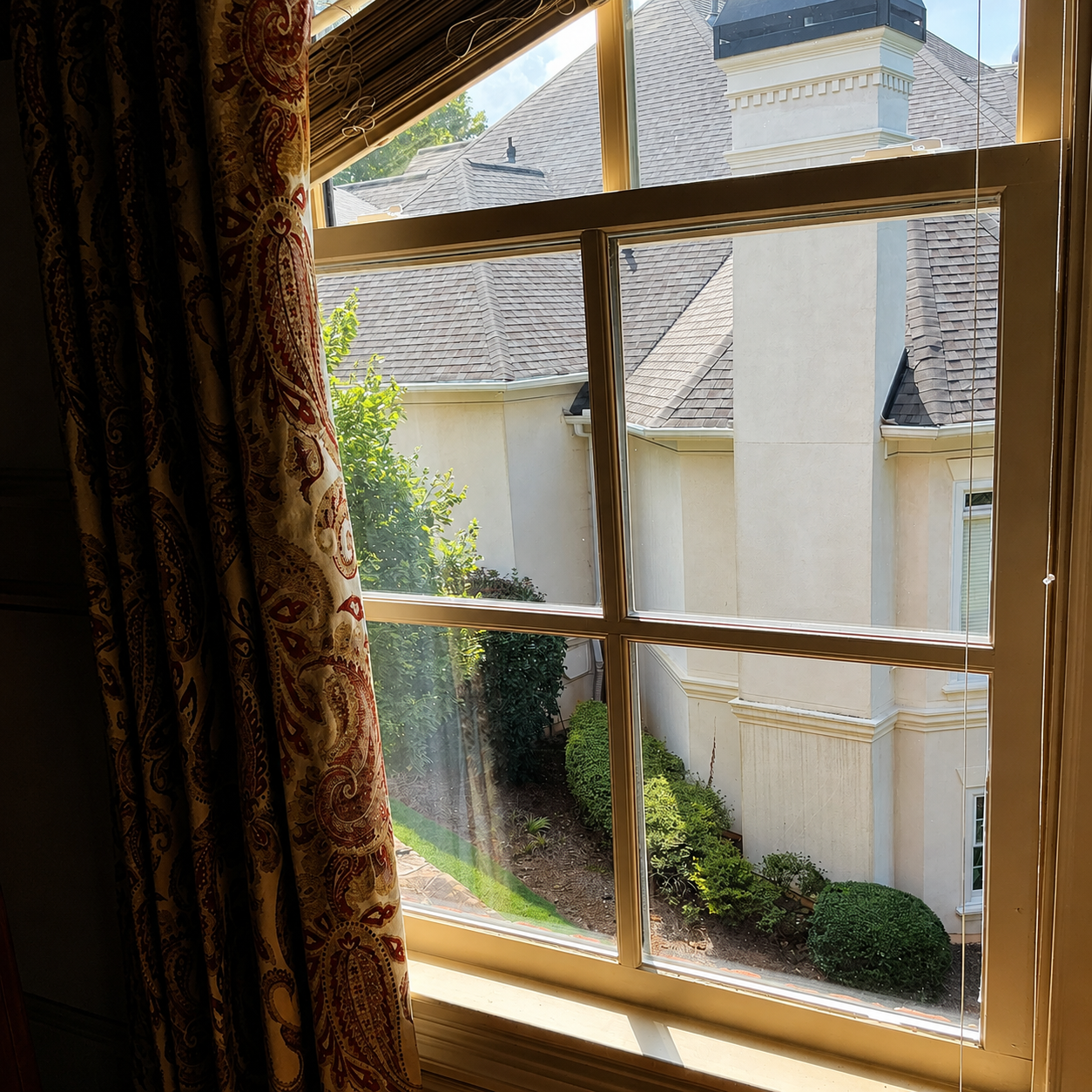 View through a window with lace curtains to a sunlit courtyard and white houses.