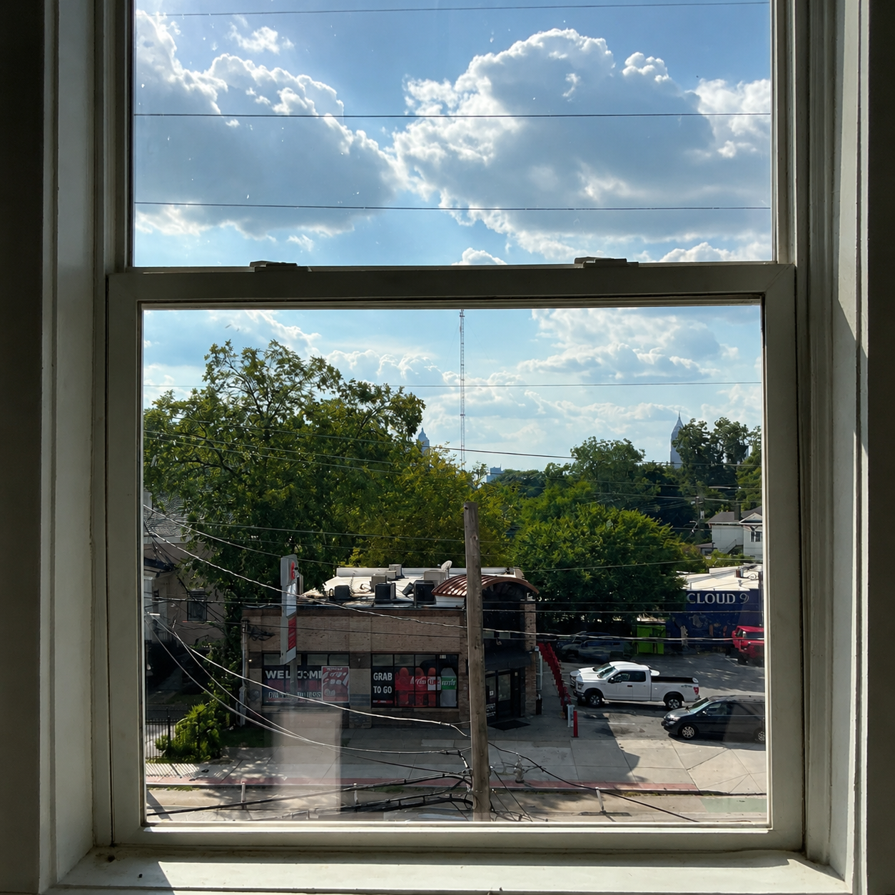 View through a window of a sunny street with trees, parked cars, and clouds overhead