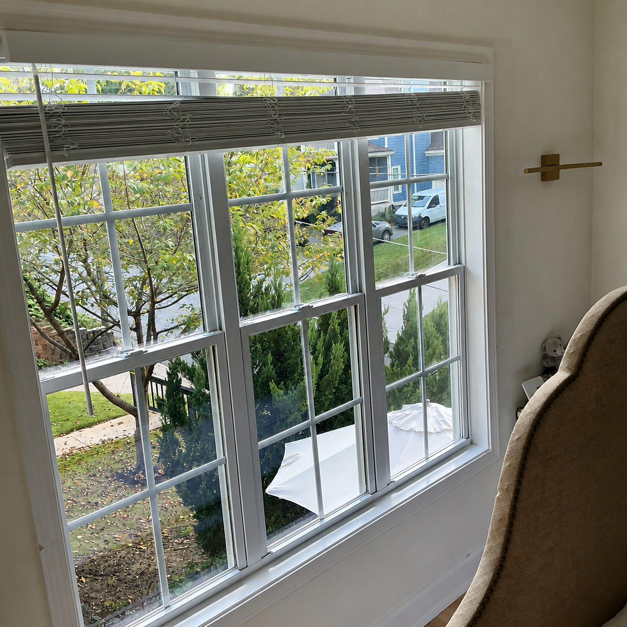 Sunny window with white blinds, looking out onto trees and houses, beside a beige chair.