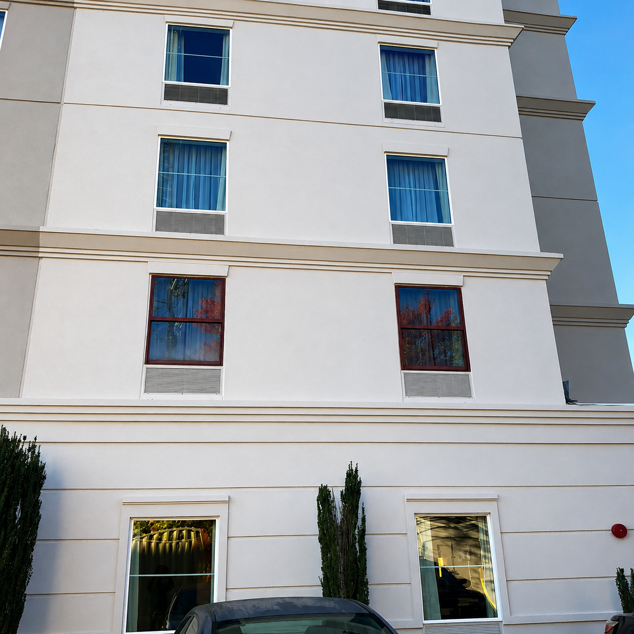 White multi-story building facade with blue windows and a few ground-floor doors and shrubs