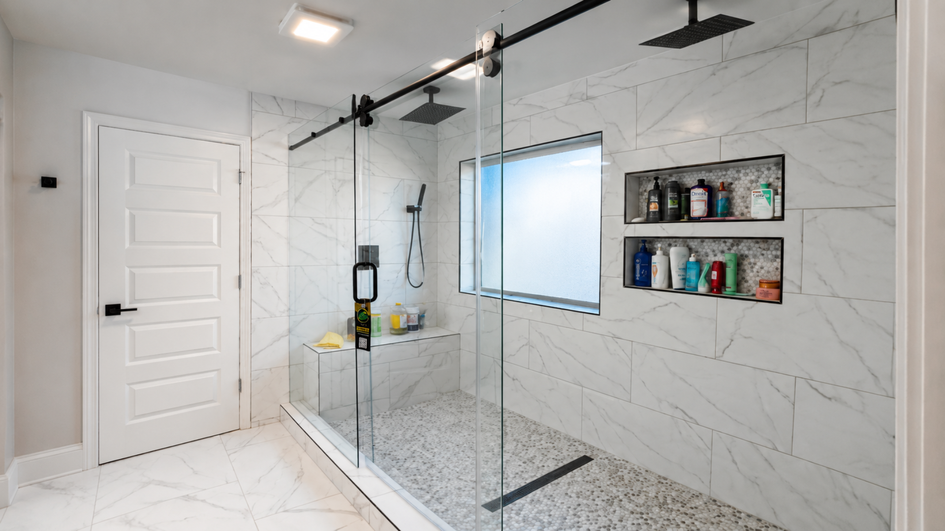 Modern shower with glass enclosure, white marble-look tile, black fixtures, and wall-mounted shampoo shelves.
