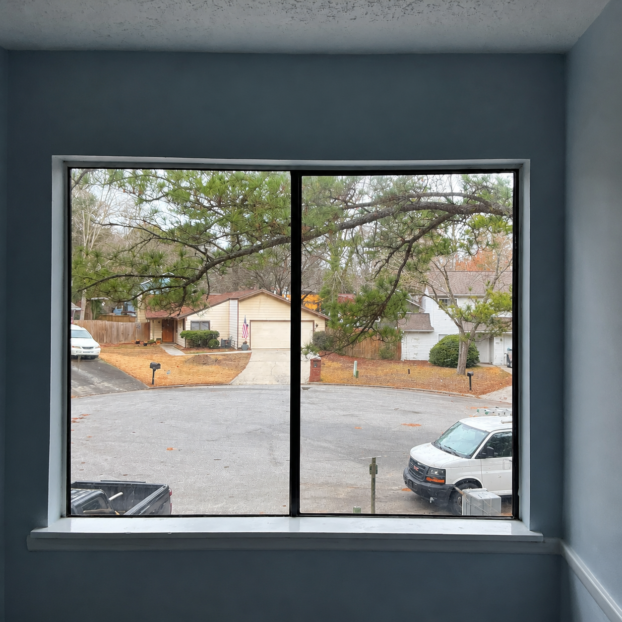View through a window of a quiet suburban street with trees, houses, and parked cars.