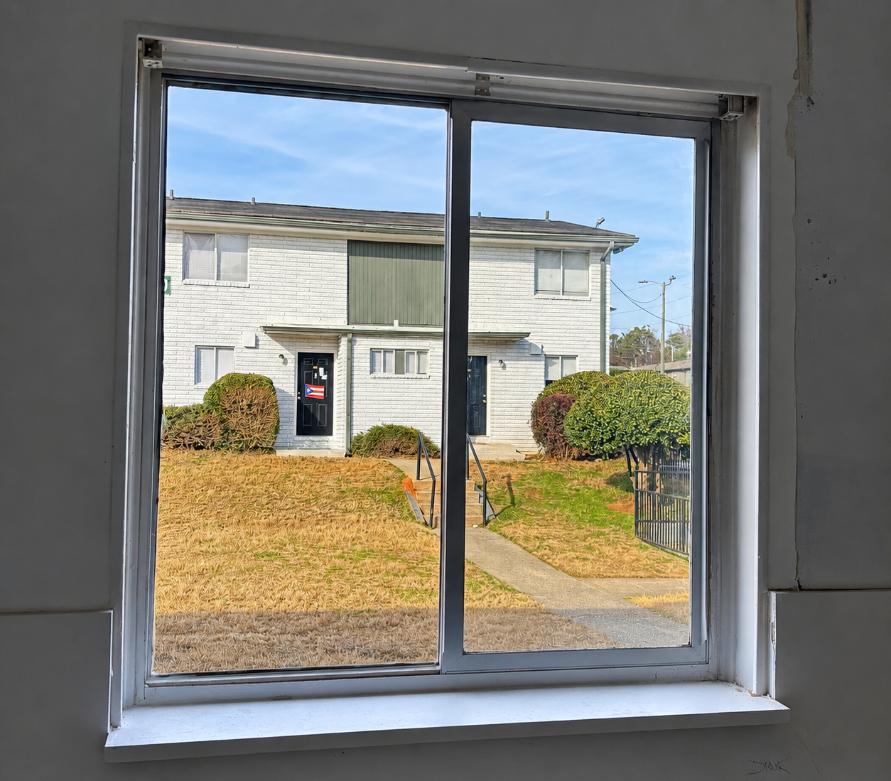 View through a window to a two-story white apartment building and autumn-colored shrubs outside.