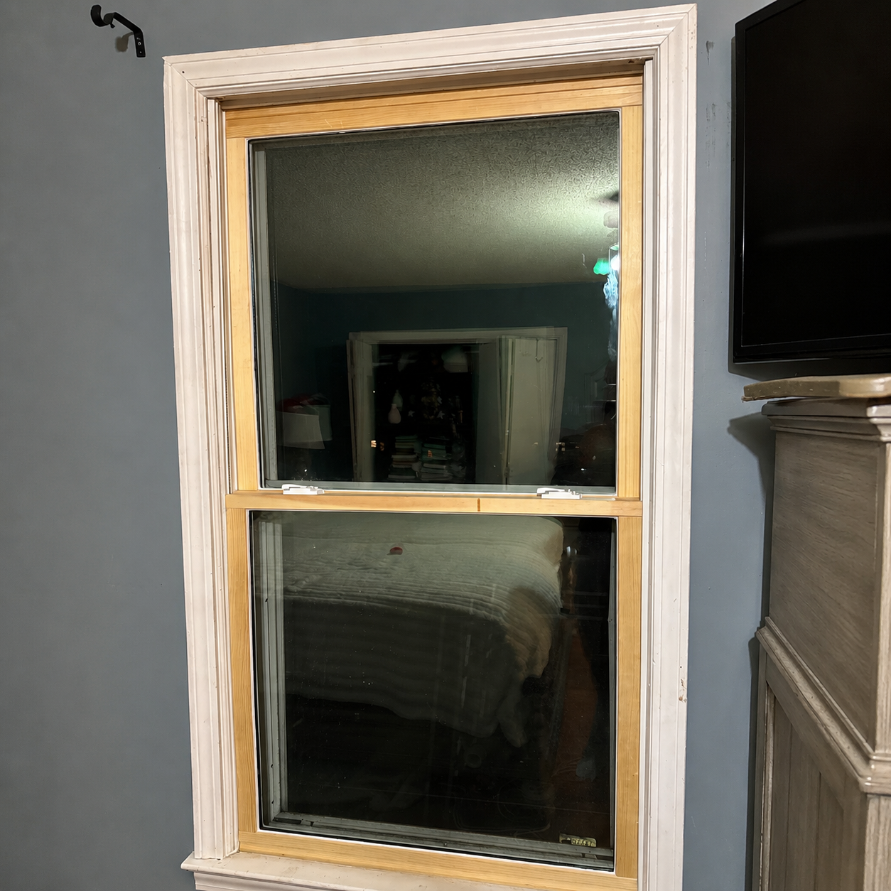Wood-framed window at night with dark reflections inside a gray room beside a TV and fireplace mantel