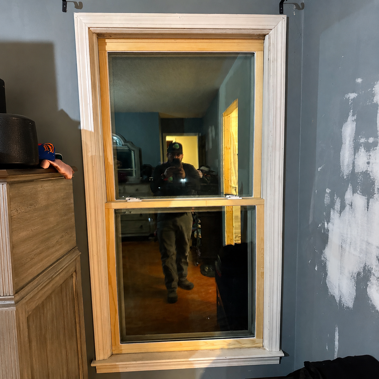 Framed wall mirror reflecting a person in a dim hallway beside blue-gray walls and a wooden cabinet