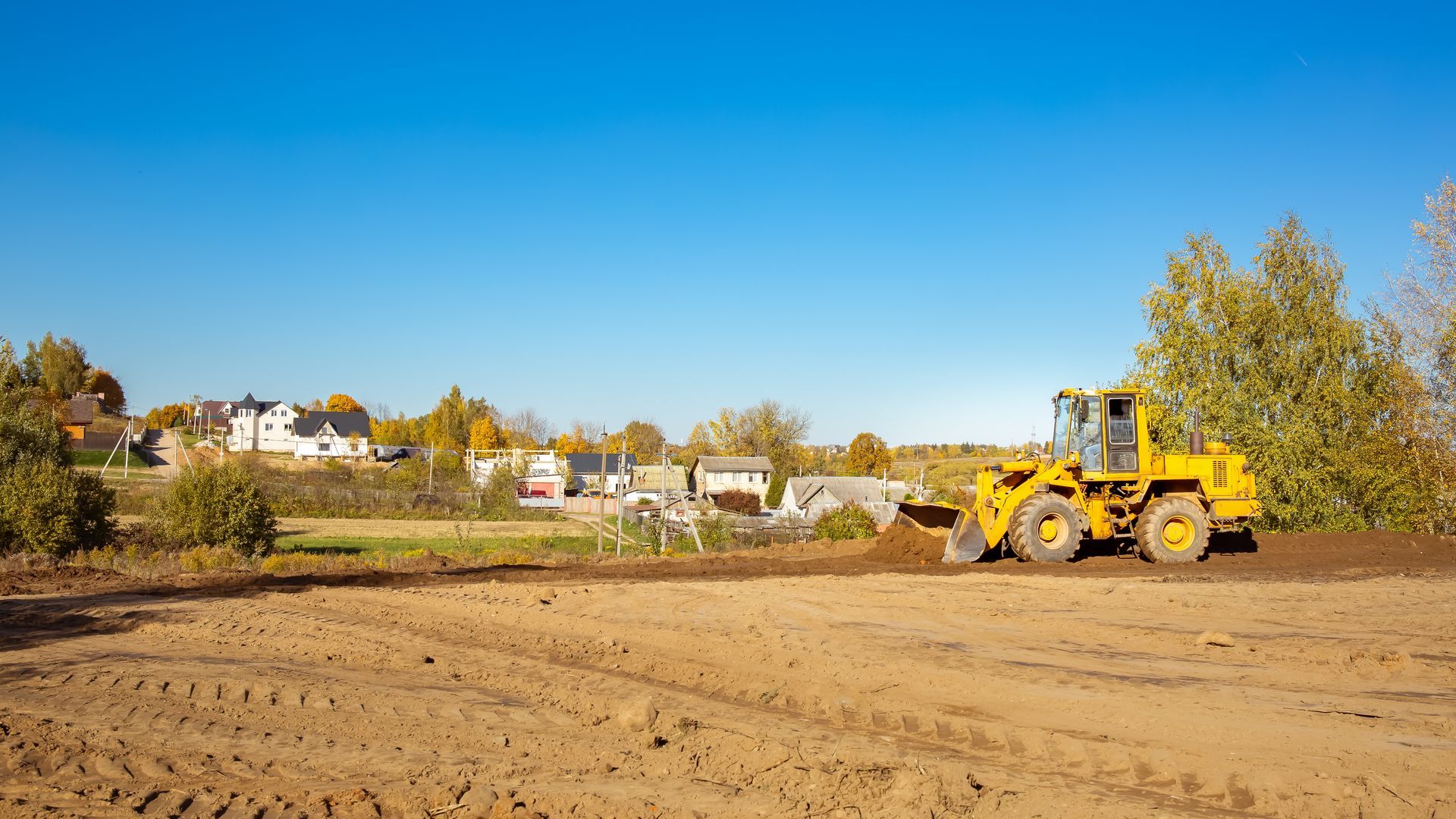 Yellow construction loader on a dirt site with houses and autumn trees under a clear blue sky