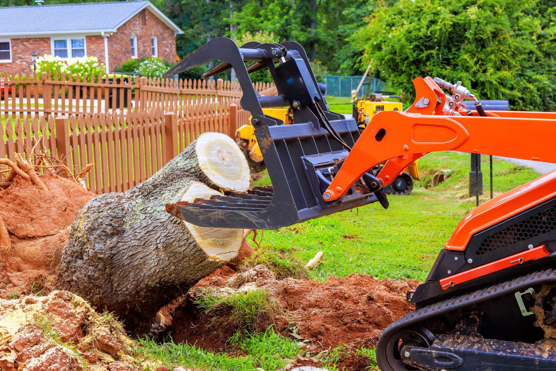Mini excavator pulling a large tree stump in a muddy yard near a house and fence