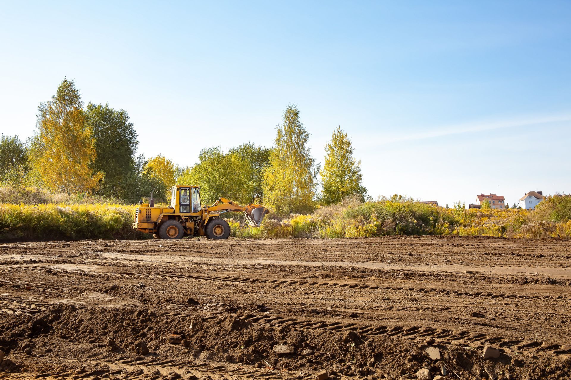 Yellow tractor plowing a field beside trees under a clear blue sky