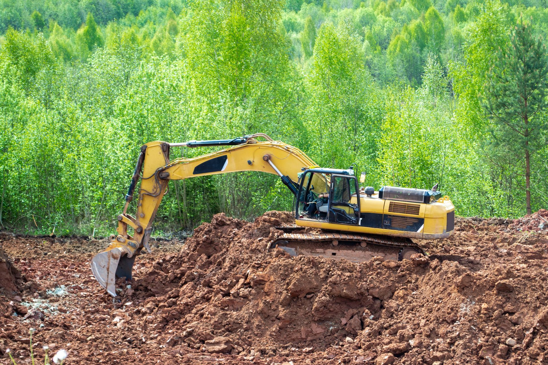 Yellow excavator digging earth on a muddy construction site with green forest in the background