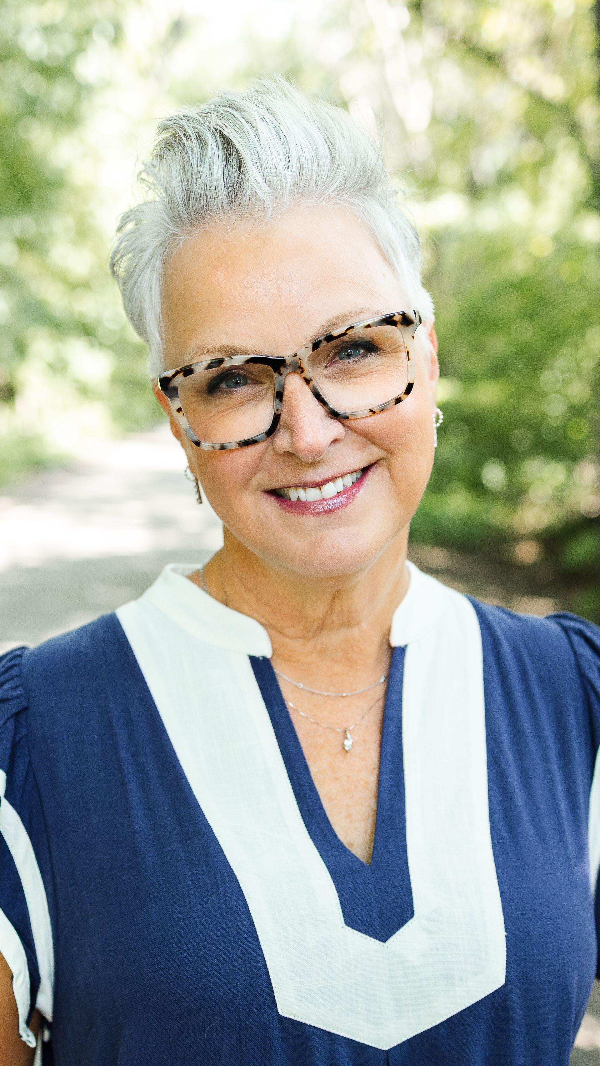 Woman with short gray hair and glasses smiles. Wearing a striped shirt and necklace, standing against a gray backdrop.