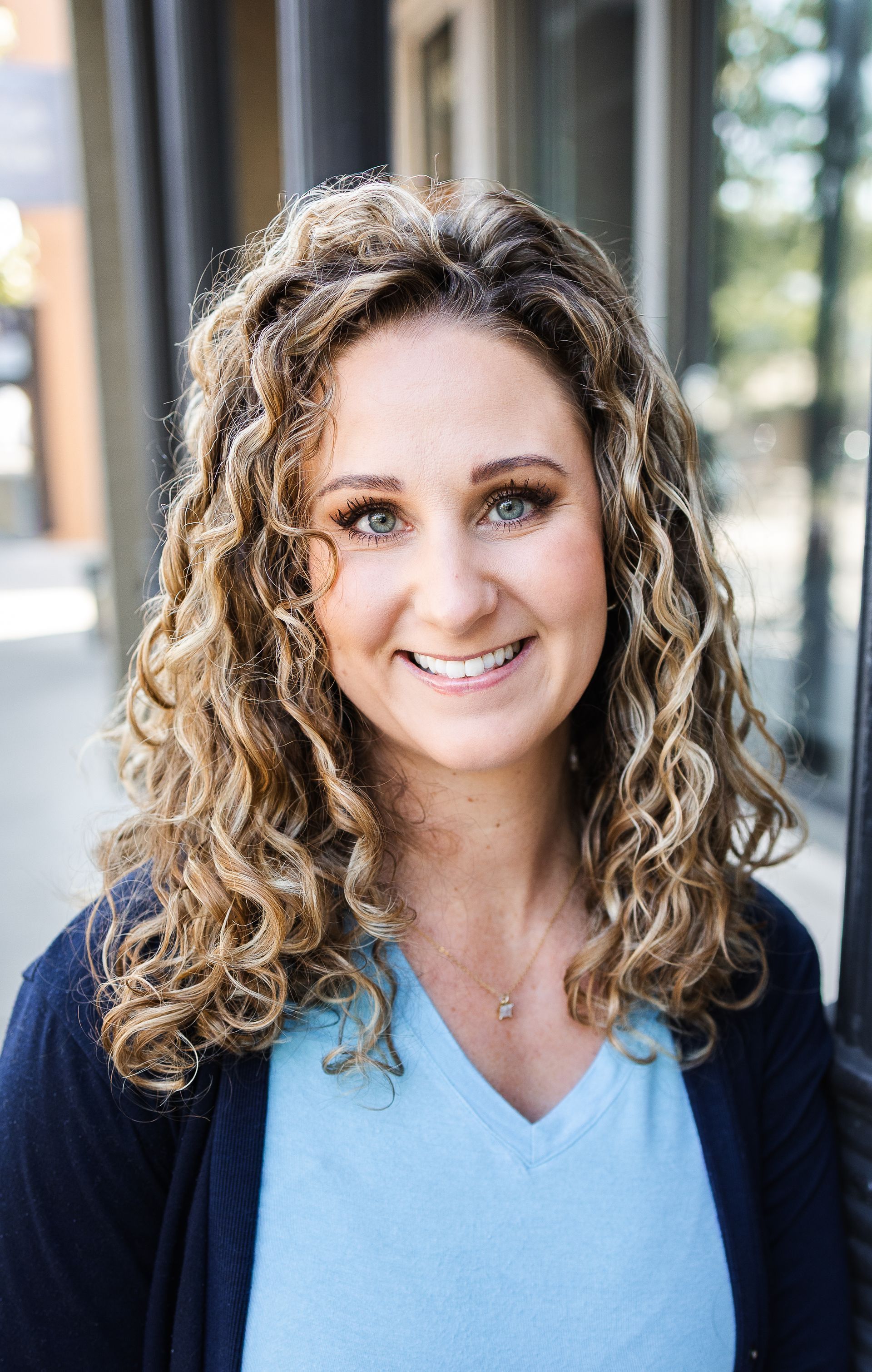 Woman with curly brown hair, smiling, wearing a blue top and grey blazer.
