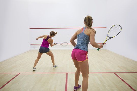Two women play racquetball on an indoor court, one in pink shorts swinging at the ball.
