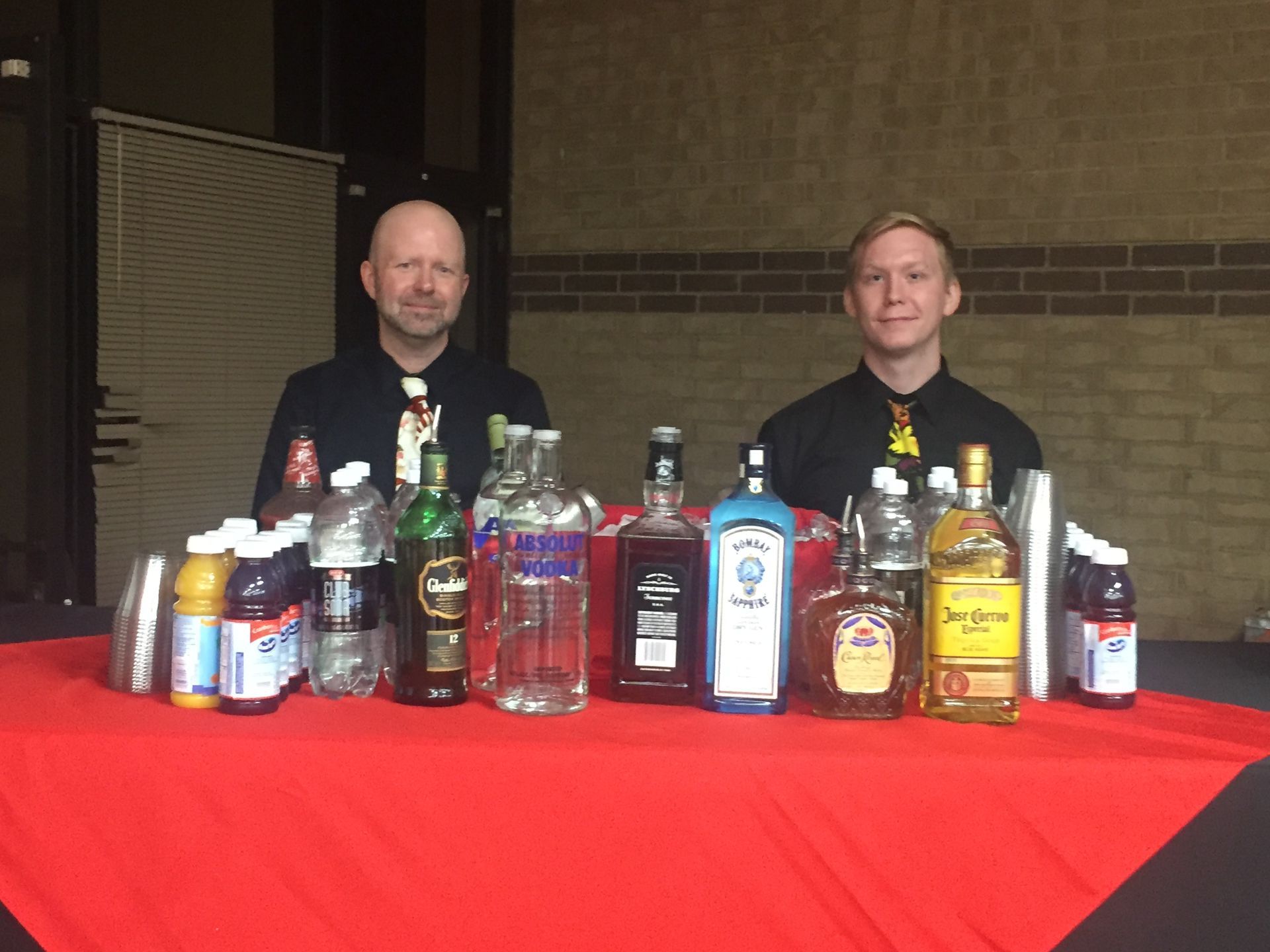 Two men are standing in front of a table filled with bottles of alcohol.