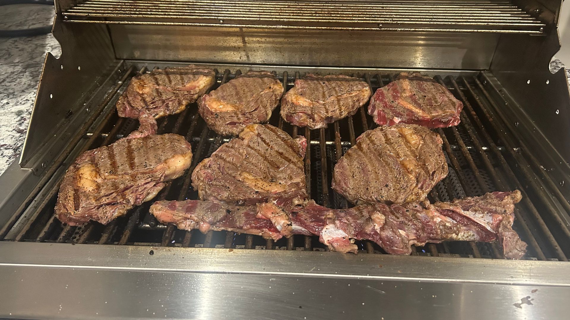 Steaks and bacon are cooking on a grill.