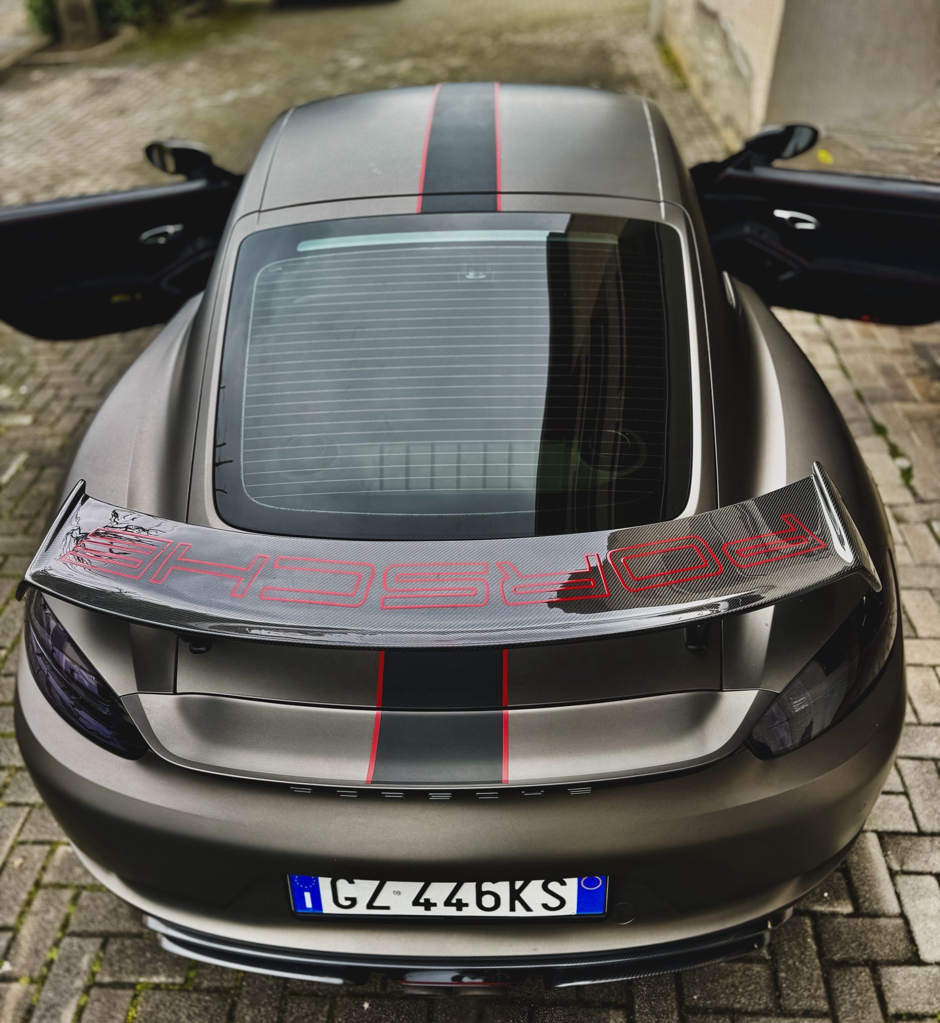 Auto sportiva grigio opaco con striscia rossa e spoiler posteriore rialzato parcheggiata su un vialetto