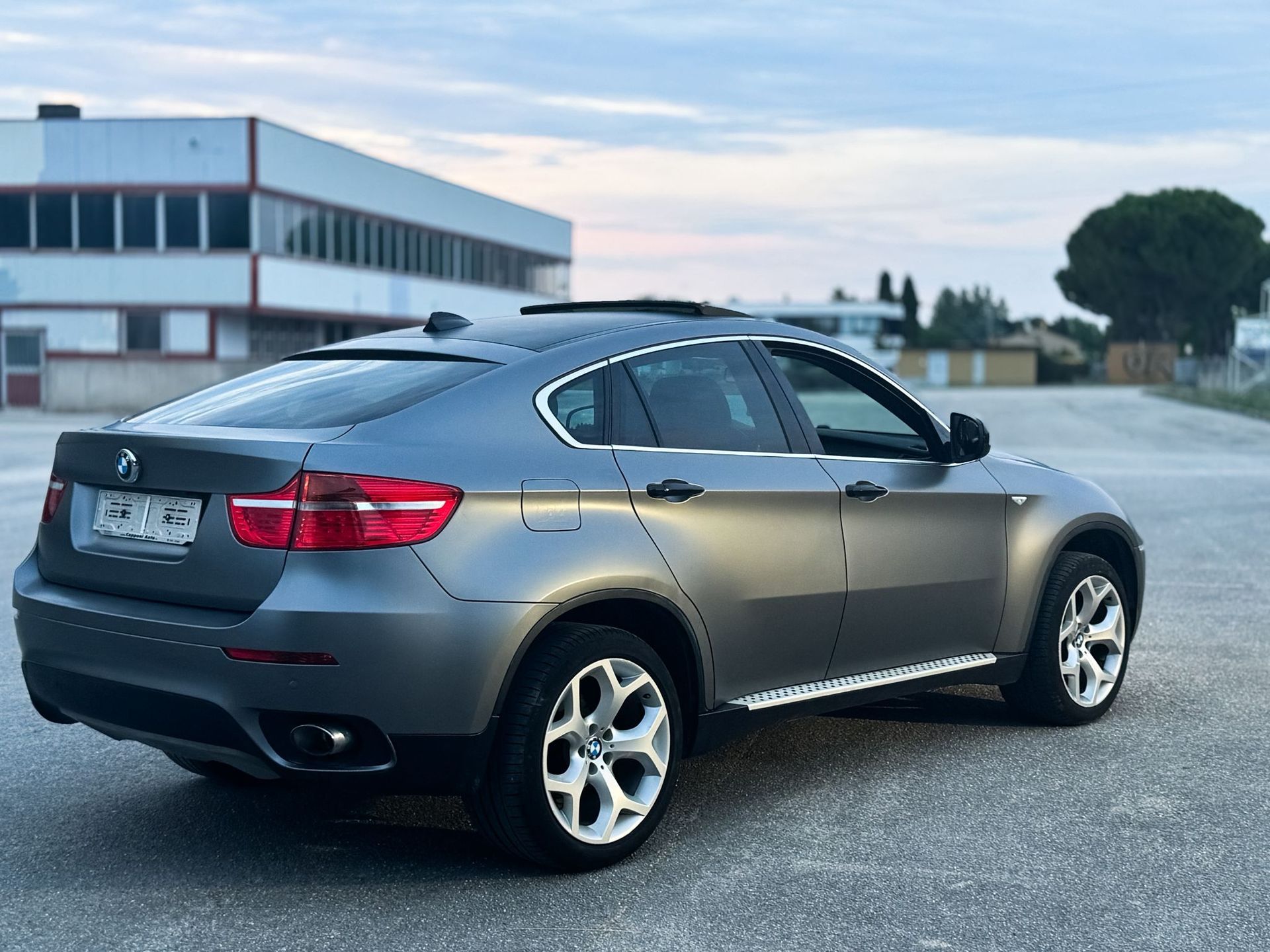 Un SUV BMW grigio parcheggiato sull'asfalto vicino a un edificio moderno al crepuscolo.