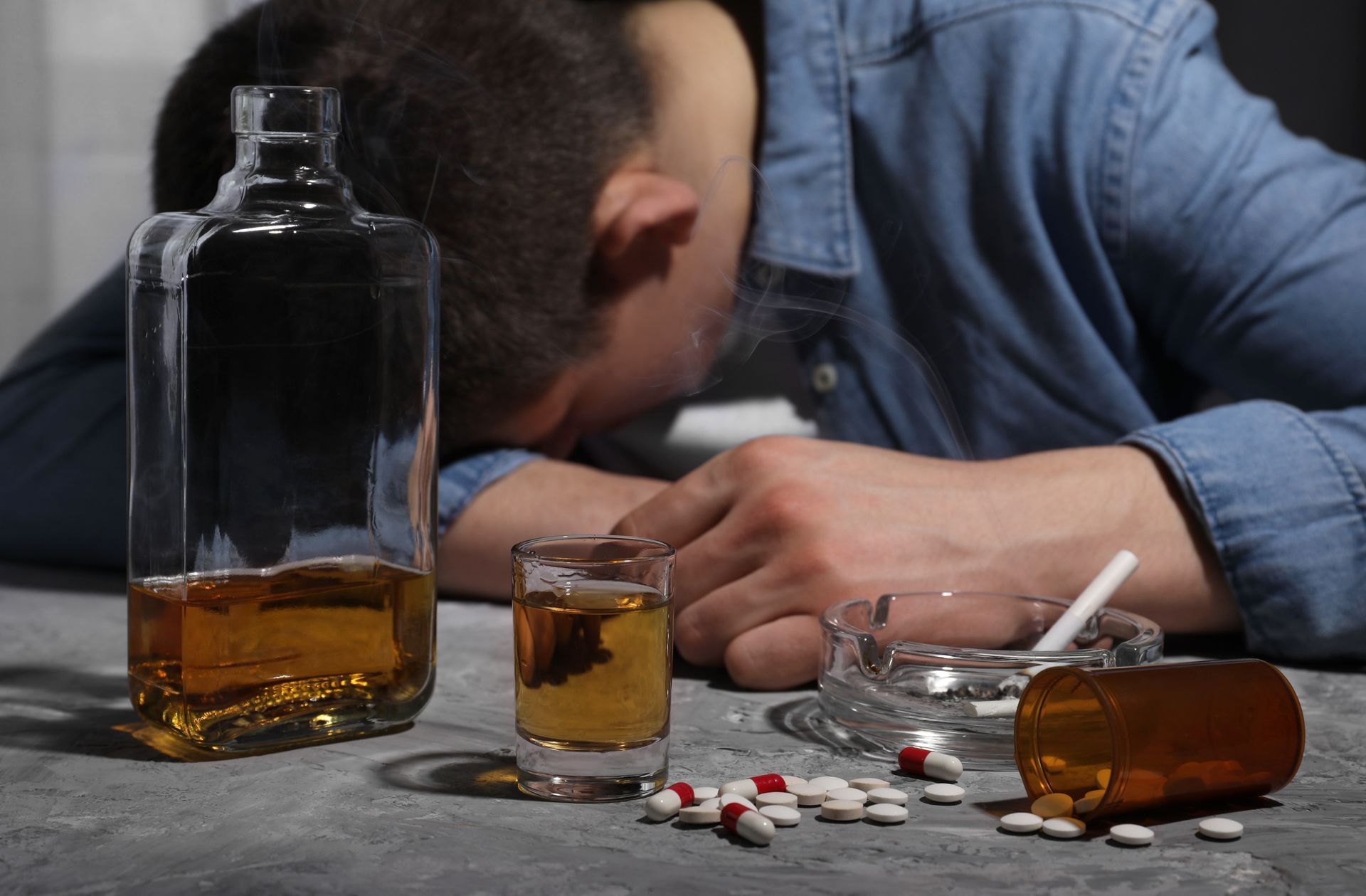 Man with head down beside alcohol bottles, pills, and an ashtray on the floor