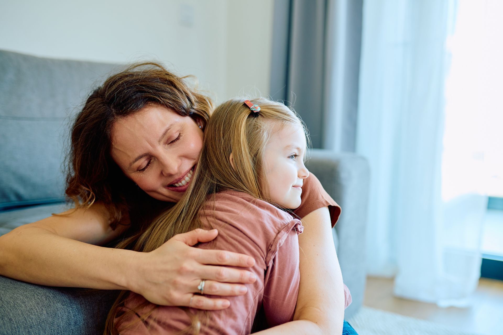 Woman hugging a young child on a couch in a bright room, both with eyes closed.