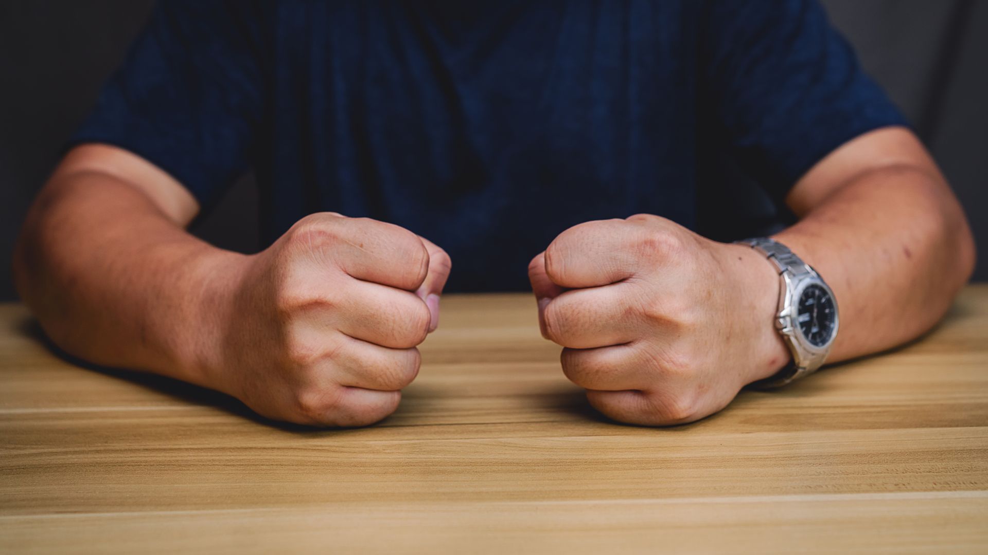 Clenched fists resting on a wooden table, with a person in a dark shirt and a wristwatch visible.