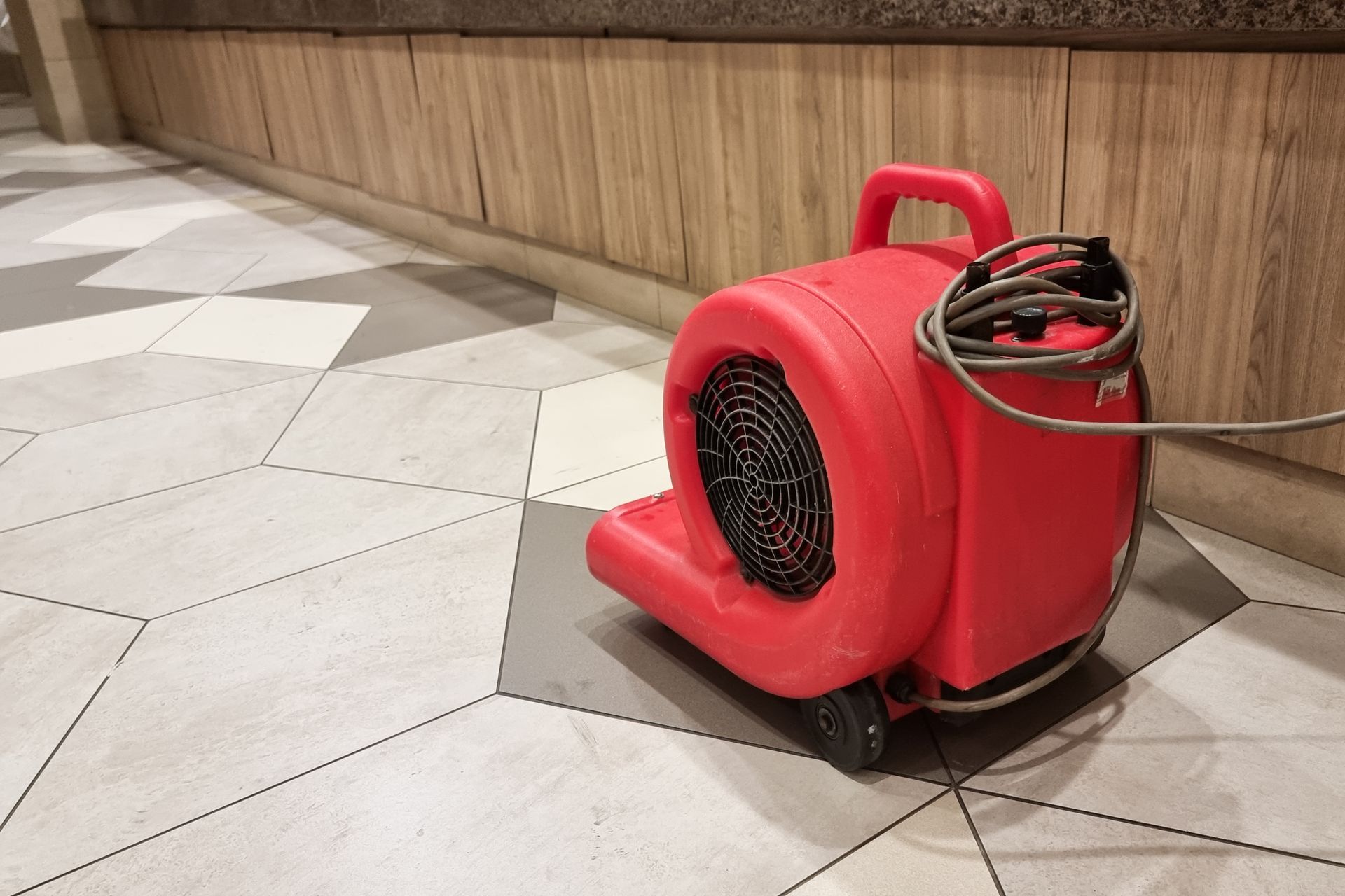 Red industrial floor fan with coiled cord on a tiled floor beside a wood-paneled wall.