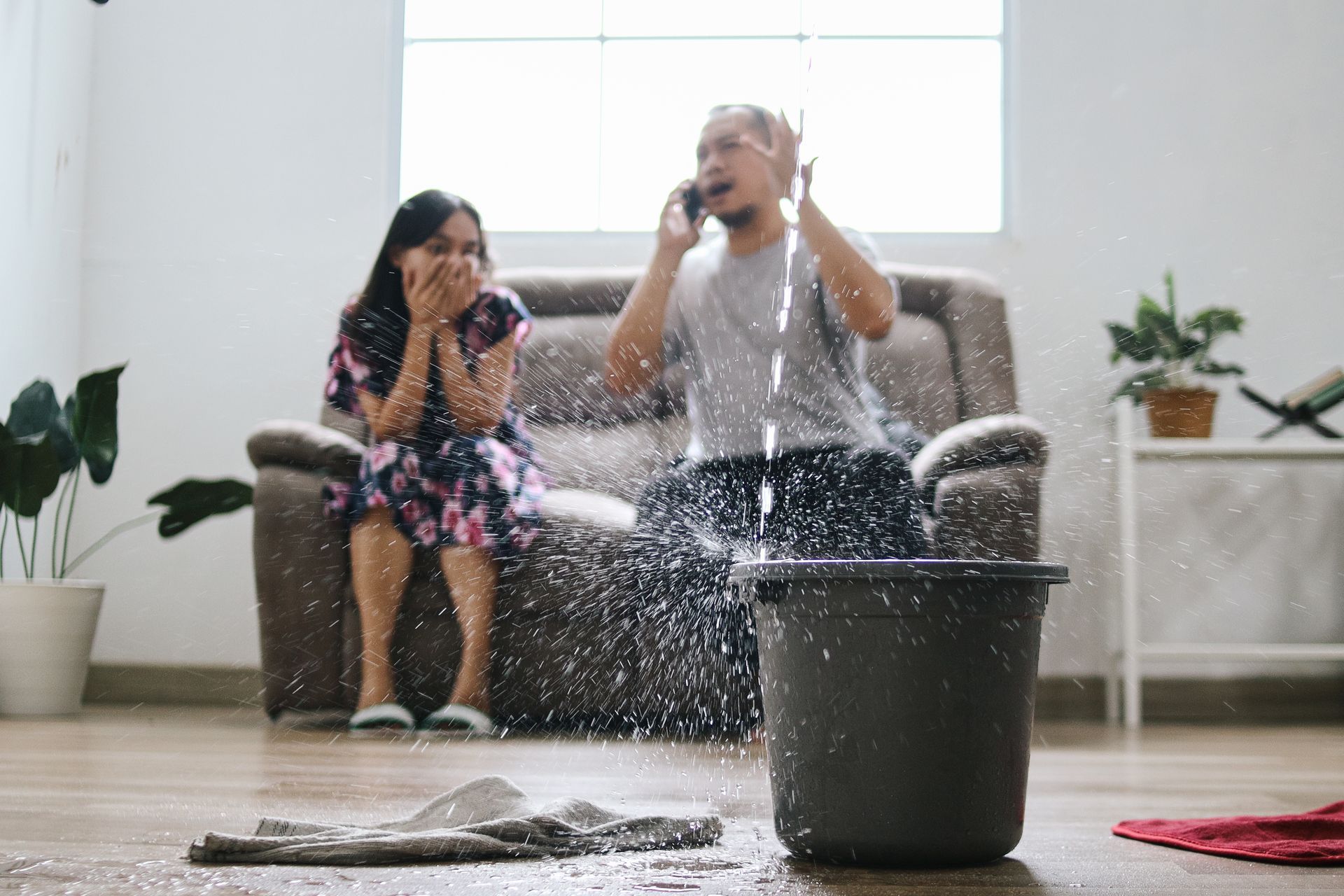 Man on phone, reacting to water splashing from ceiling, next to a bucket. Woman on couch shocked.